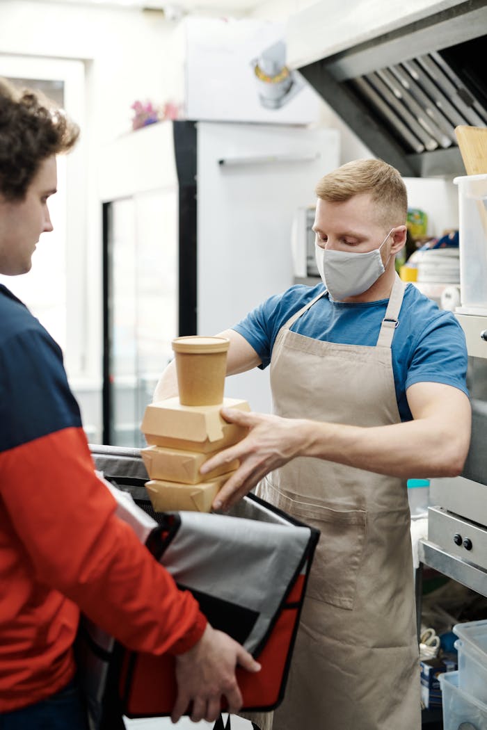 Masked worker packs takeaway food for delivery in restaurant kitchen.